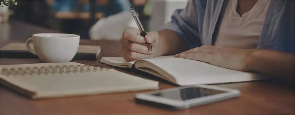 A person writing in a notebook with a cup of coffee.