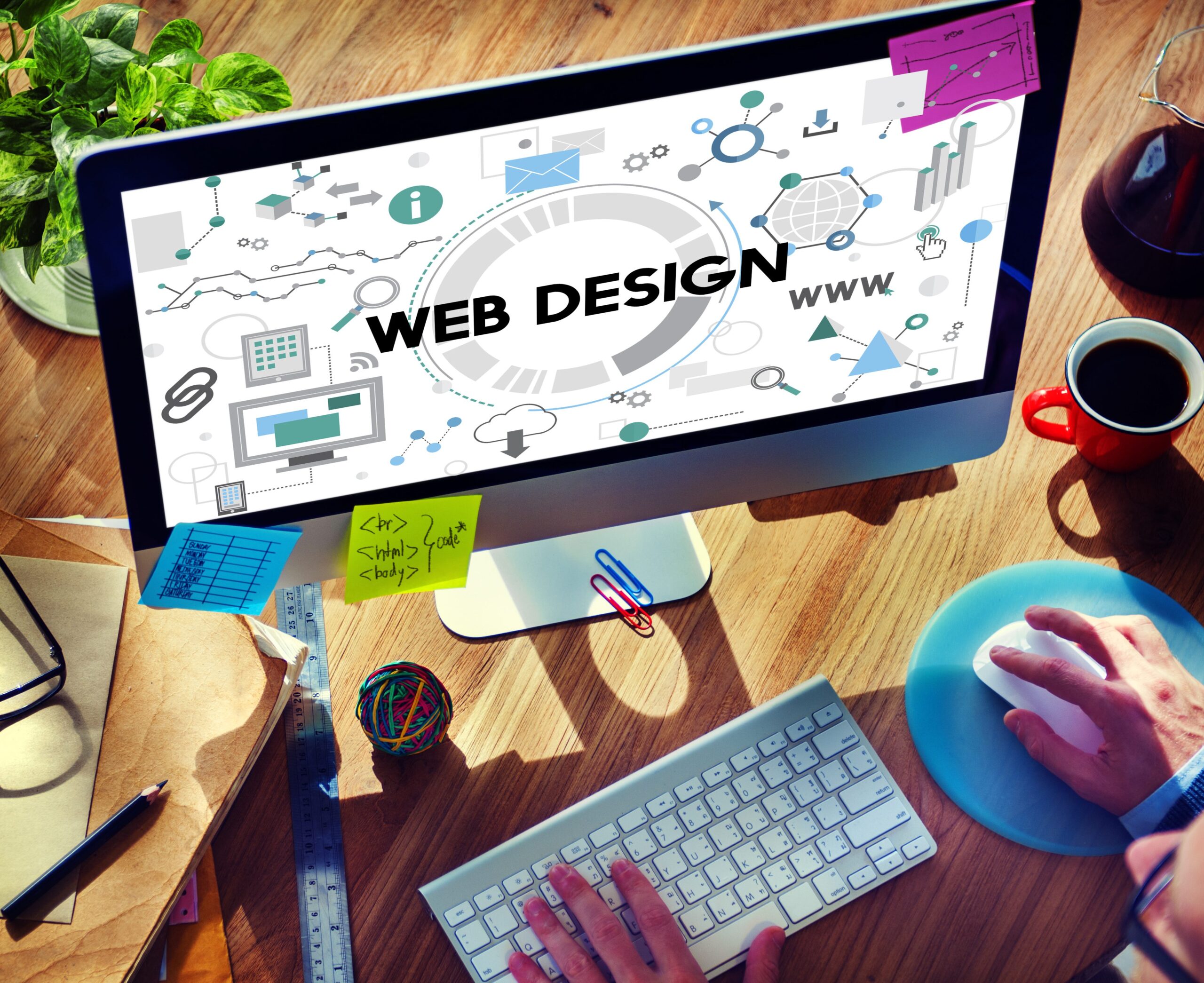 Computer screen displaying "Web Design" with graphics, surrounded by office supplies. Person using mouse and keyboard.