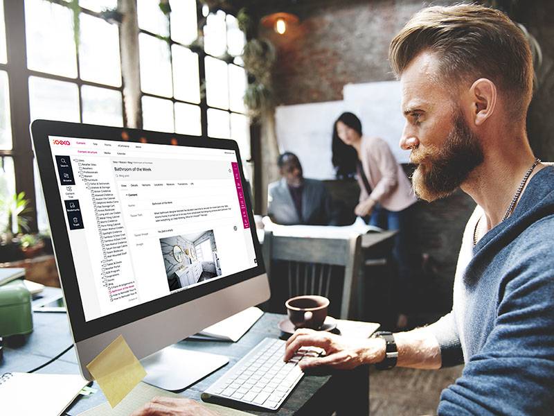Man with a beard works at a desktop computer in a modern office, while two colleagues collaborate in the background near windows.