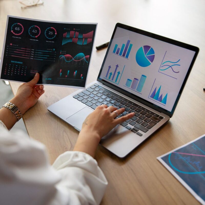 Person analyzing graphs and charts on a laptop and printed paper, with various data visualizations displayed on both screen and documents.