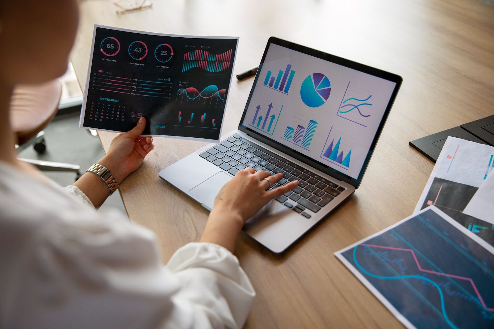 Person analyzing graphs and charts on a laptop and printed paper, with various data visualizations displayed on both screen and documents.
