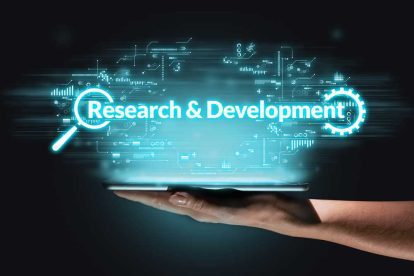 A hand holds a digital tablet displaying the words "Research & Development" with futuristic graphics and technology icons in the background.