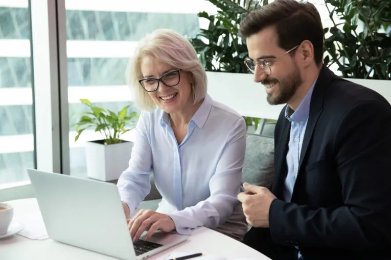 Two colleagues, a woman and a man, sit at a desk and look at a laptop screen together in a modern office with large windows and plants.