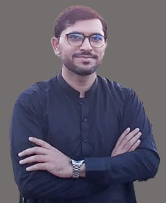A man with short hair, glasses, and a beard is standing with his arms crossed, wearing a black traditional outfit and a wristwatch, against a plain gray background.