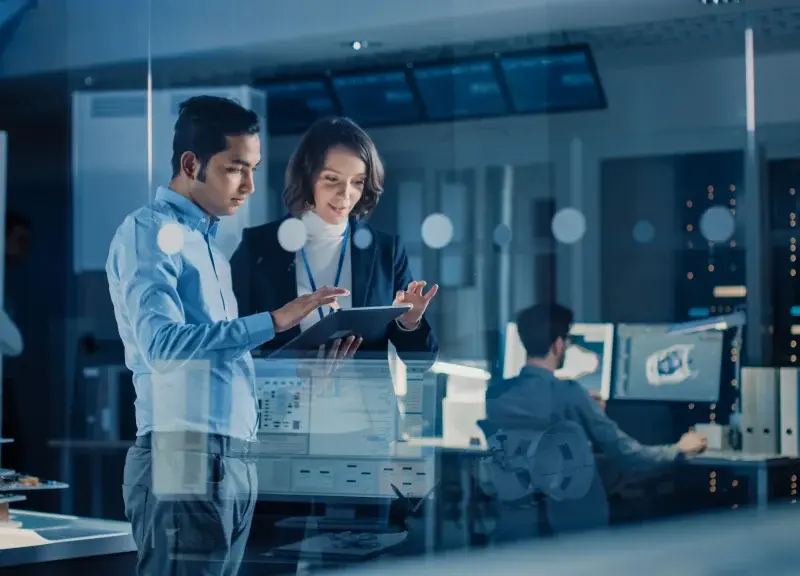 Two professionals review information on a tablet in a modern office with computer monitors and technology equipment visible in the background.