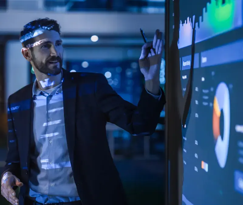 A man in a suit points at charts and graphs on a large digital screen during a presentation in a dimly lit room.