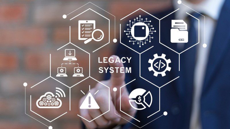 A person in a suit points at a virtual interface featuring icons related to technology and data, with the words "LEGACY SYSTEM" in the center.