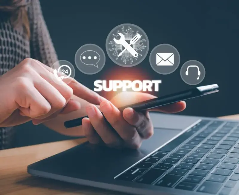 Person using a smartphone near a laptop with digital icons representing support, chat, email, and help services.