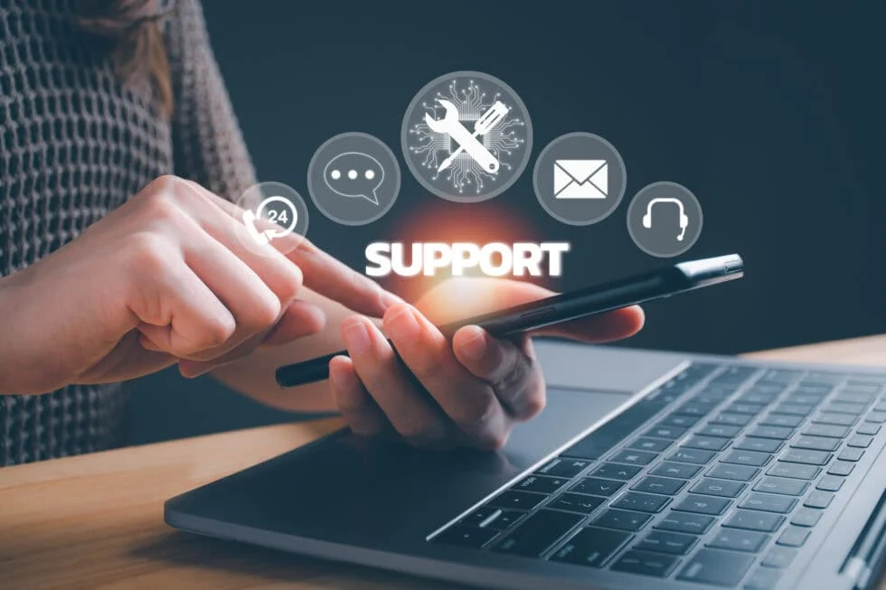 Person using a smartphone near a laptop with digital icons representing support, chat, email, and help services.
