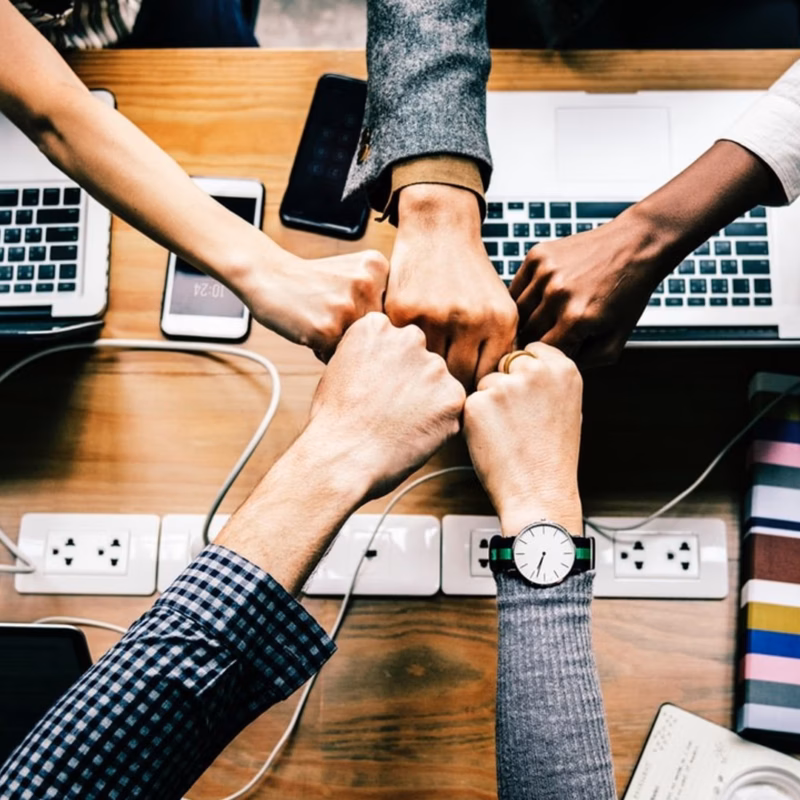 Five people seated at a table with laptops, phones, and chargers, join their fists together over the center of the table in a gesture of teamwork.