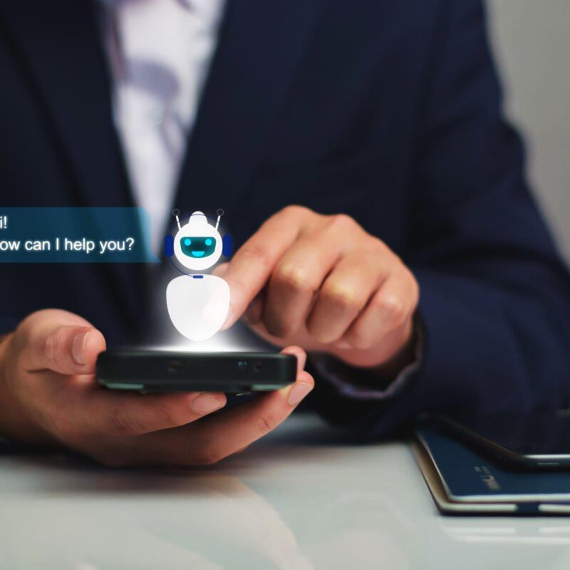 A person in a suit uses a smartphone displaying a chatbot hologram with the message, "Hi! How can I help you?" on the screen.
