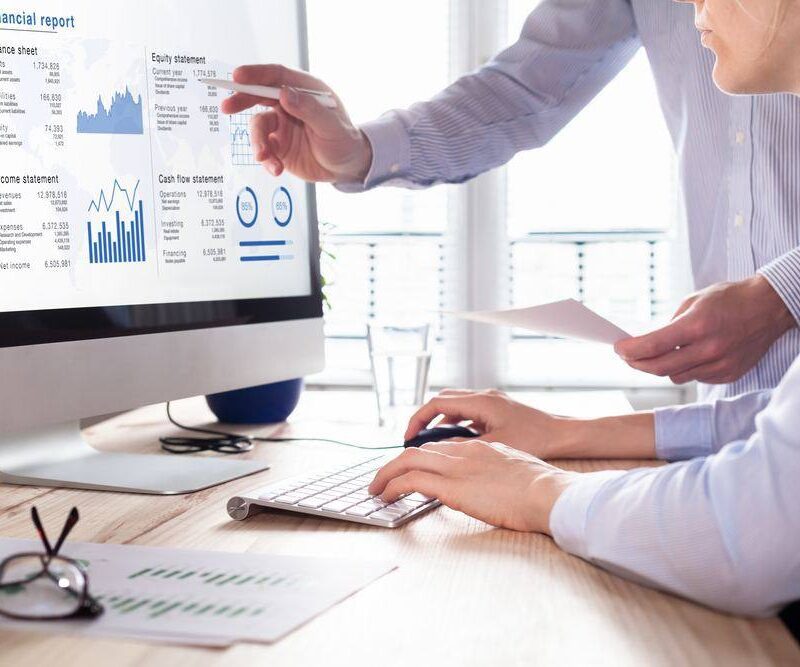 Two people review financial data on a computer screen displaying charts and statements, with printed documents and a keyboard on the desk.