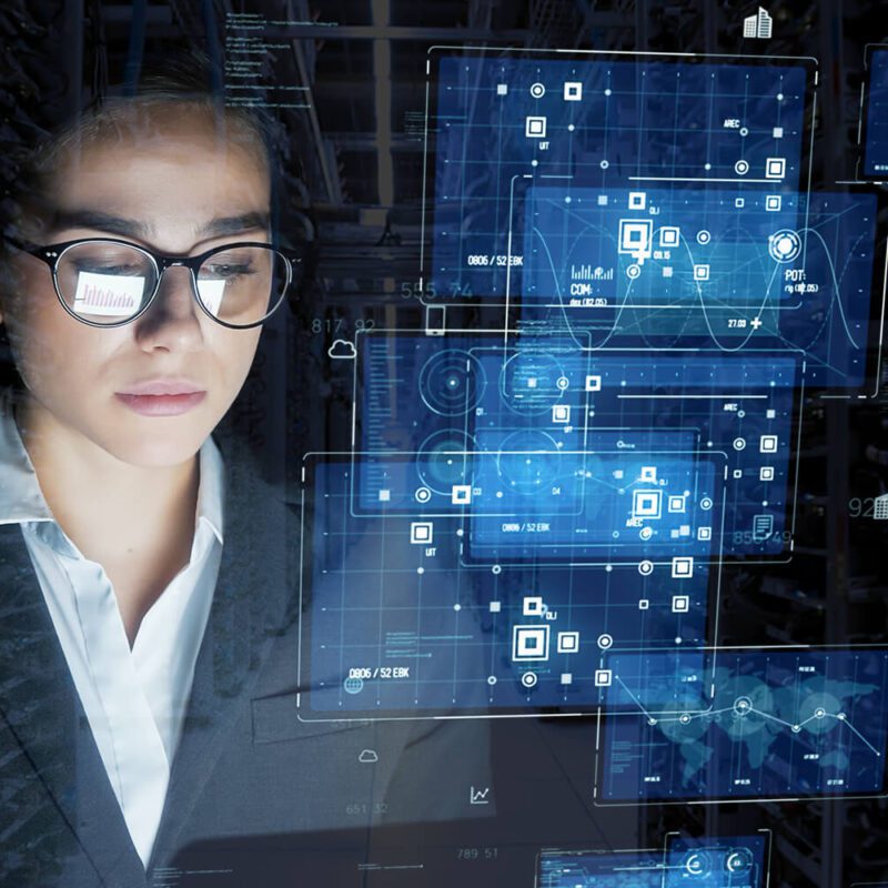 A woman in business attire looks at virtual digital interfaces displaying data, charts, and network graphics in a technology-driven environment.