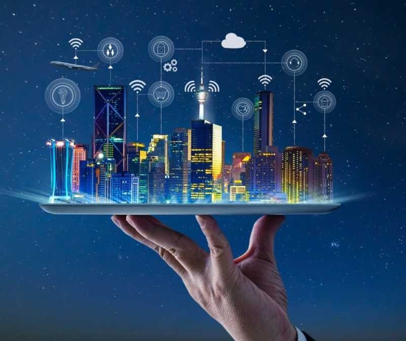 A hand holds a tablet displaying a city skyline with digital icons above buildings, representing smart city technology and connectivity.