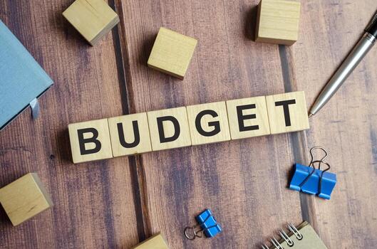 Wooden blocks spell out “BUDGET” on a wooden surface, surrounded by a pen, binder clips, and other blocks.