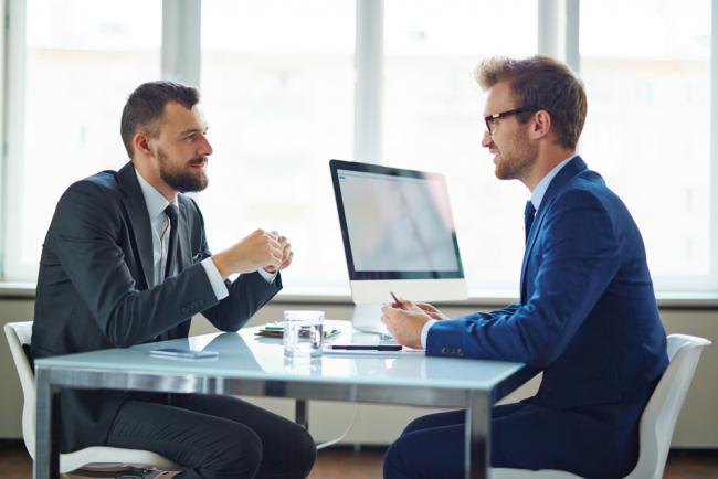Two men in business suits sit across from each other at a glass desk in an office, having a discussion with a computer monitor between them.