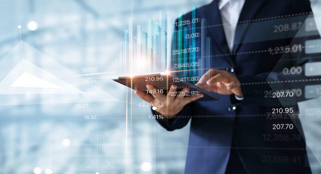 A person in a suit uses a tablet with digital financial graphs and stock prices overlayed in the background.