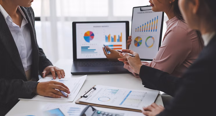Three people in business attire discuss financial charts and graphs displayed on a laptop screen and paper documents during a meeting at a desk.