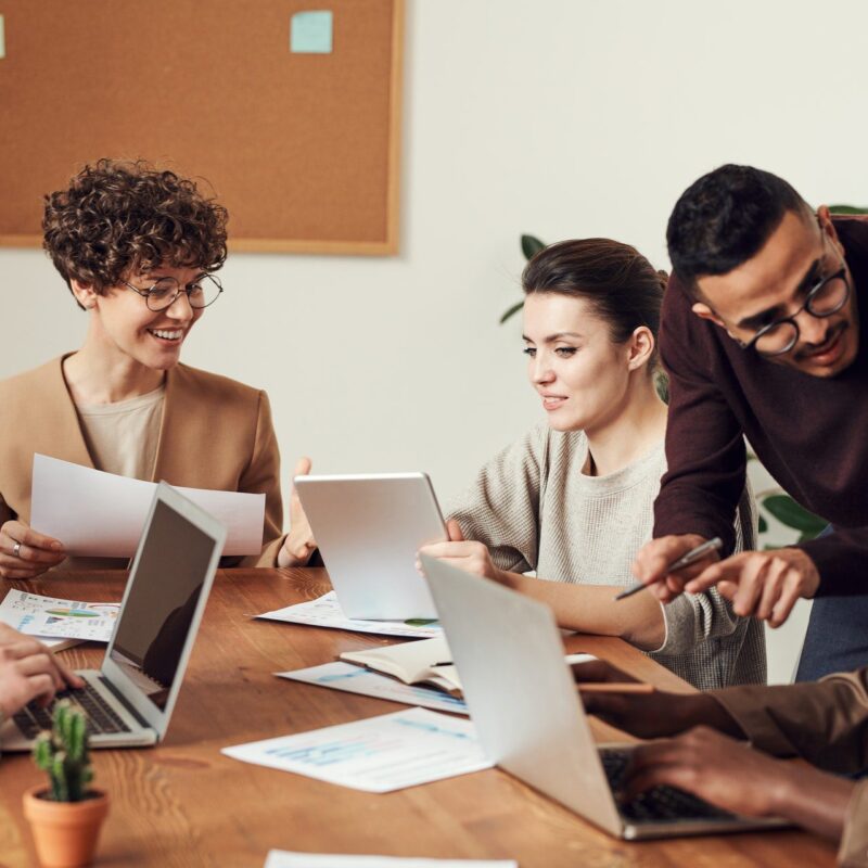 Five people sit at a table with laptops and papers, discussing and collaborating in an office setting with a corkboard in the background.