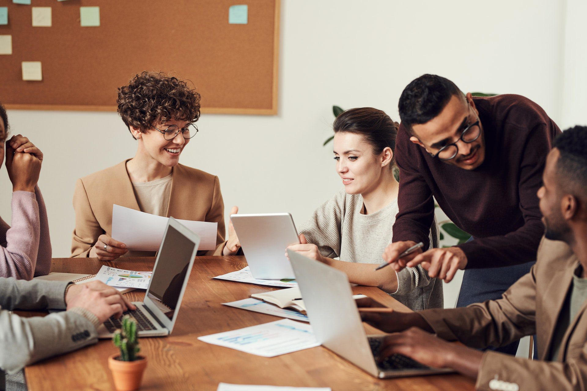 Five people sit at a table with laptops and papers, discussing and collaborating in an office setting with a corkboard in the background.
