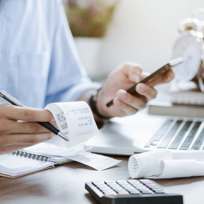 A person reviews receipts and uses a smartphone while working at a desk with a laptop, calculator, notepad, and clock.