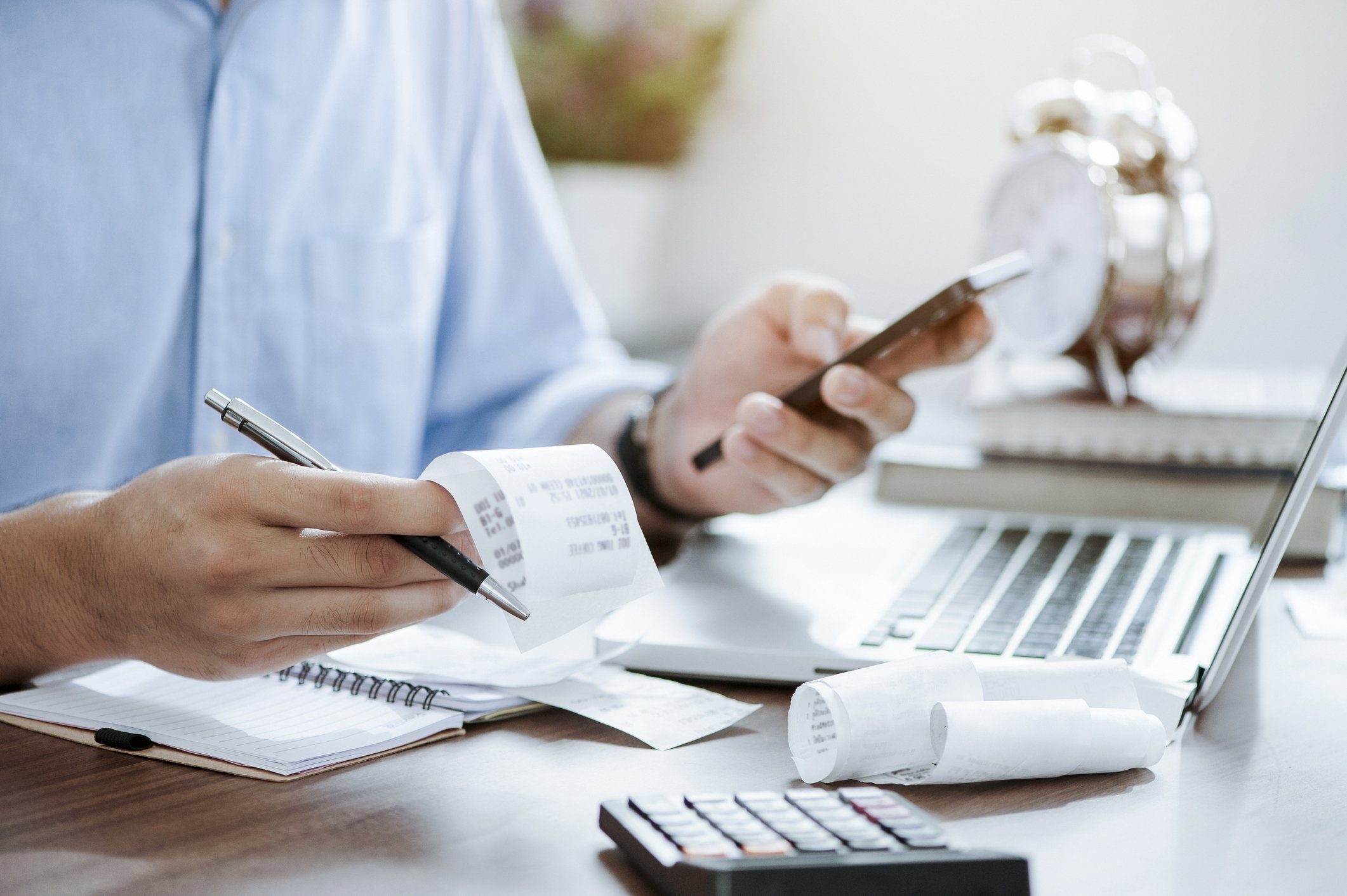 A person reviews receipts and uses a smartphone while working at a desk with a laptop, calculator, notepad, and clock.