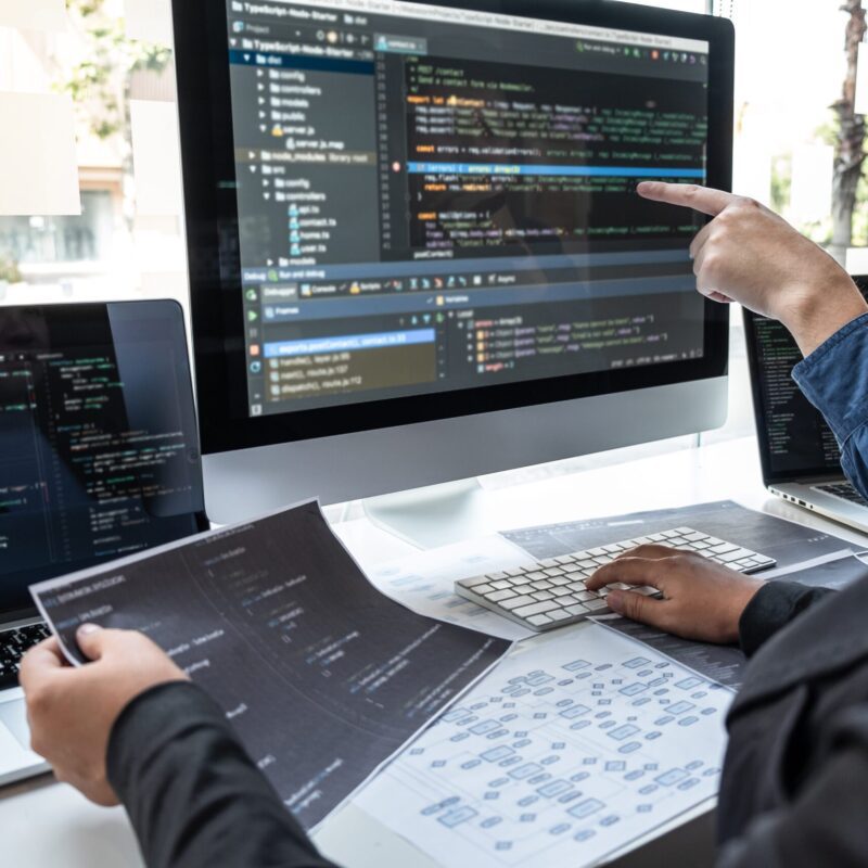 Two people working at a desk with computer monitors displaying code, printed documents, and a keyboard, discussing software development.
