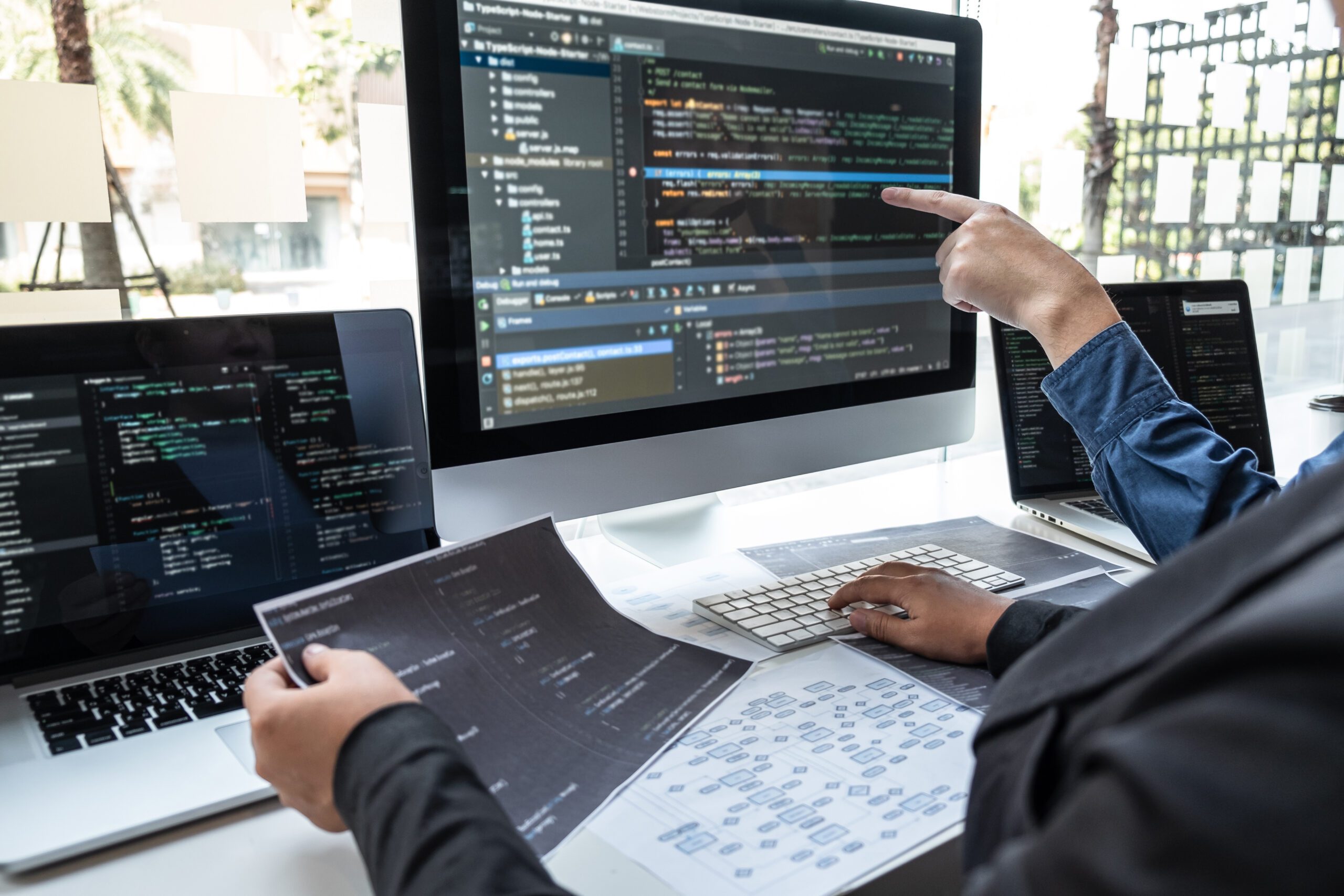 Two people working at a desk with computer monitors displaying code, printed documents, and a keyboard, discussing software development.