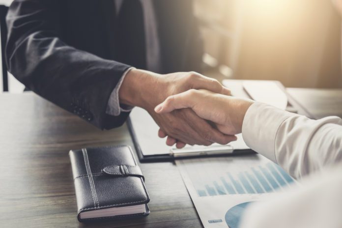 Two people in business attire shake hands over a desk with documents, a notebook, and a chart, suggesting a professional agreement or meeting.