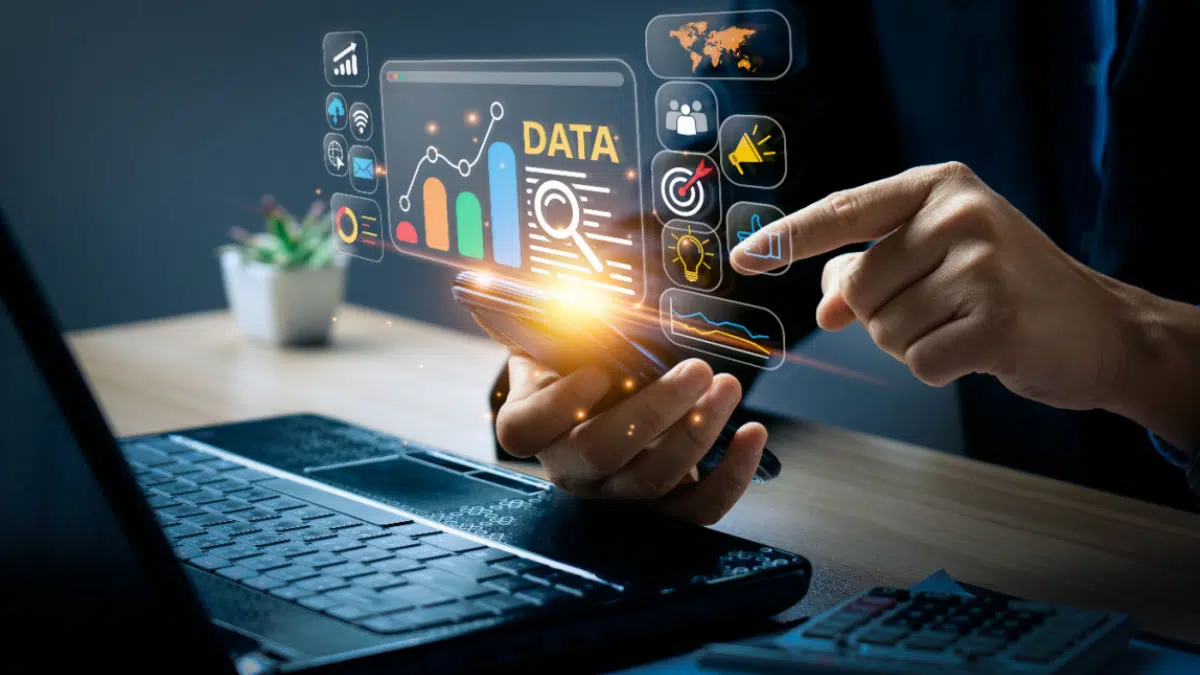 A person uses a smartphone near a laptop, interacting with floating data analytics graphics and digital icons representing charts, targets, and global connections.