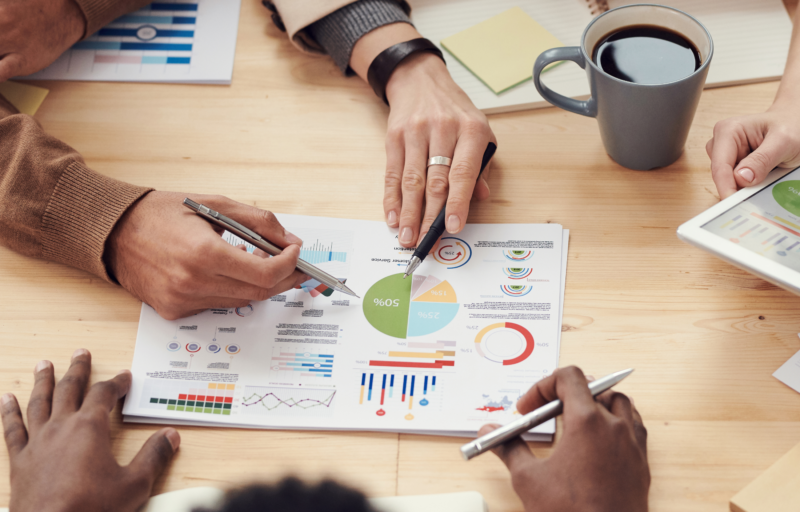 Four people sit around a table analyzing colorful business charts and graphs on paper, with hands pointing and writing, coffee mugs and a tablet nearby.
