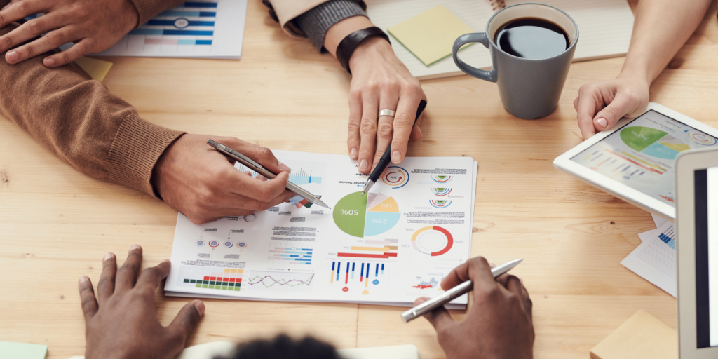 Four people sit around a table analyzing colorful business charts and graphs on paper, with hands pointing and writing, coffee mugs and a tablet nearby.
