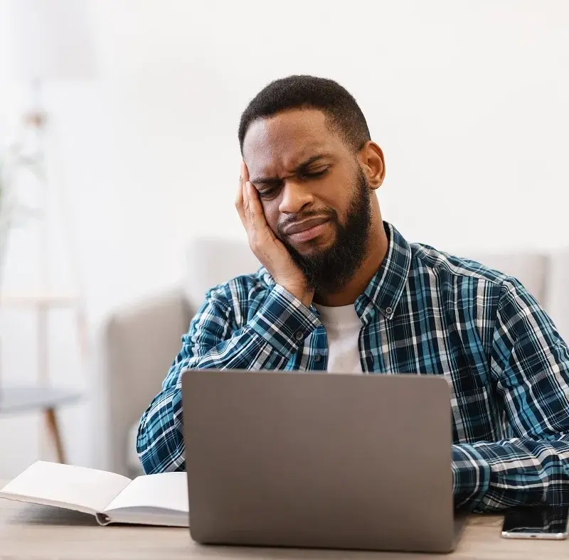 Man in plaid shirt sits at desk with laptop and notebook, holding his head and appearing to be in pain or discomfort, possibly experiencing a headache or stress.