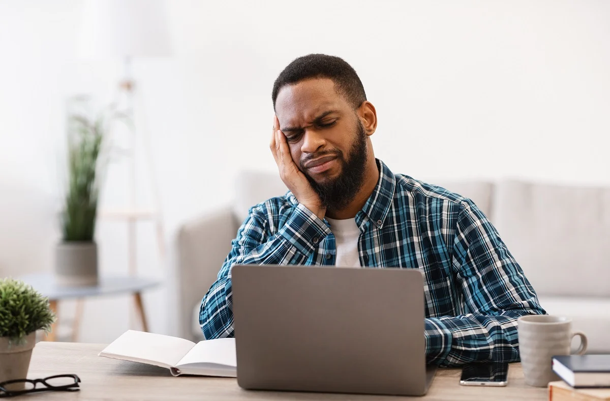 Man in plaid shirt sits at desk with laptop and notebook, holding his head and appearing to be in pain or discomfort, possibly experiencing a headache or stress.