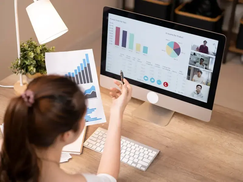 Person at desk viewing charts on paper and computer screen, participating in a video call with four people, with graphs and data visualizations displayed on the monitor.