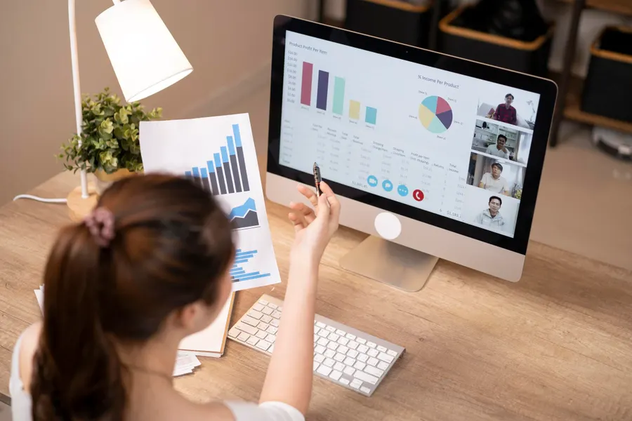Person at desk viewing charts on paper and computer screen, participating in a video call with four people, with graphs and data visualizations displayed on the monitor.