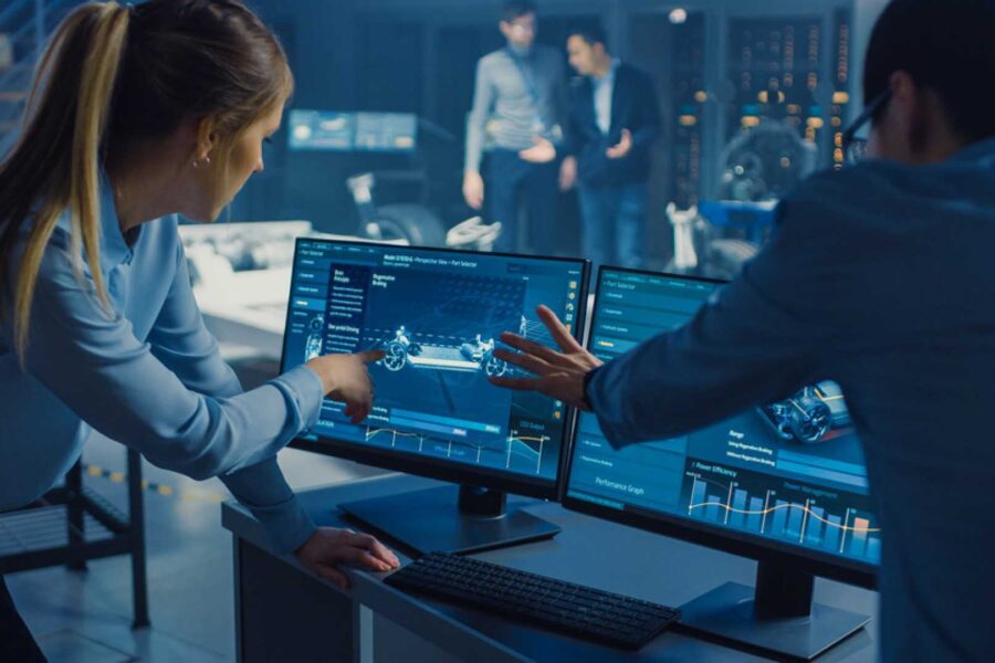 Two engineers point at computer screens displaying automotive design and performance data in a laboratory.