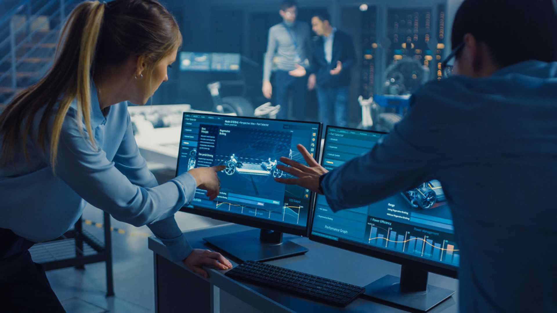 Two engineers point at computer screens displaying automotive design and performance data in a laboratory.